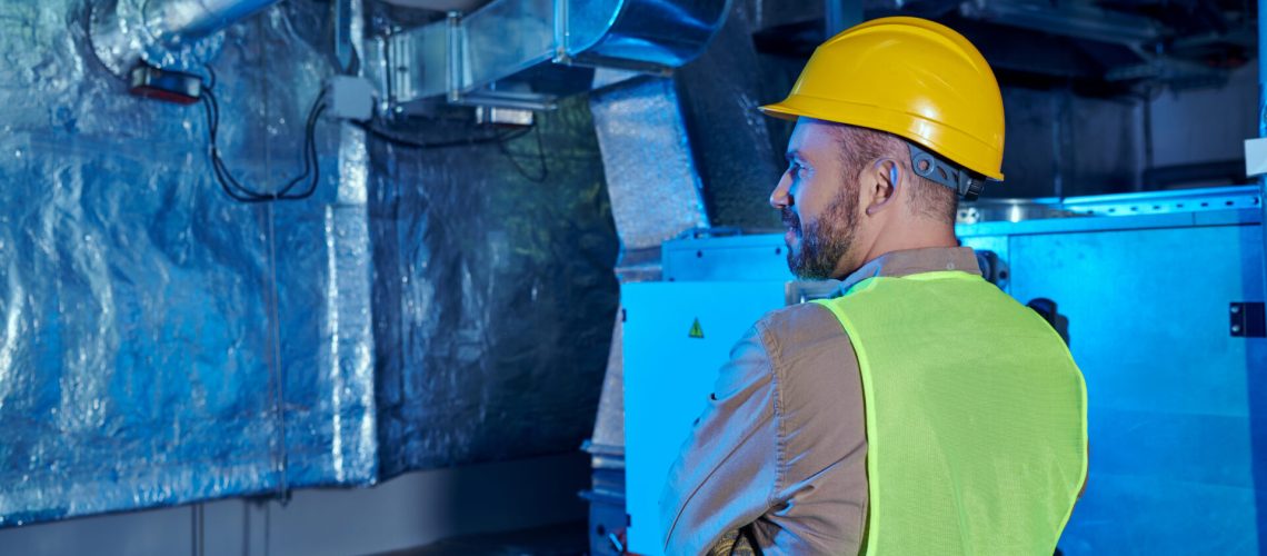 devoted jolly technician in safety vest and helmet posing with arms crossed on chest in data center